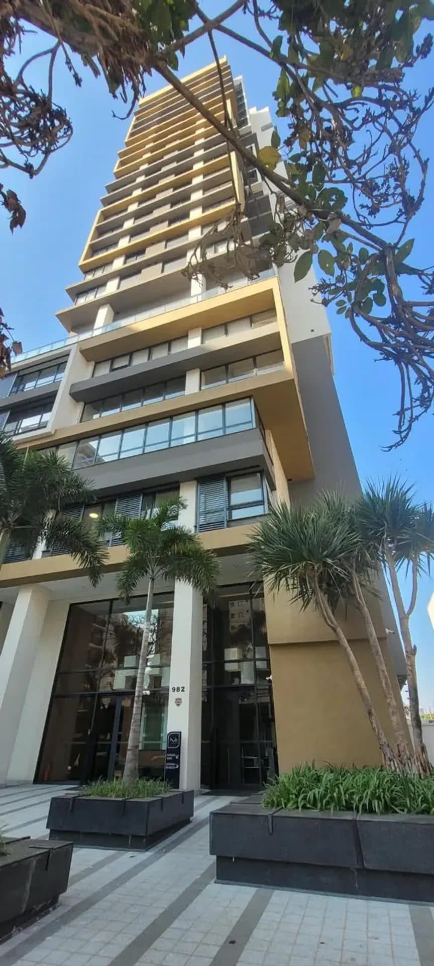 Fachada do prédio com estrutura moderna, varandas e vegetação na entrada